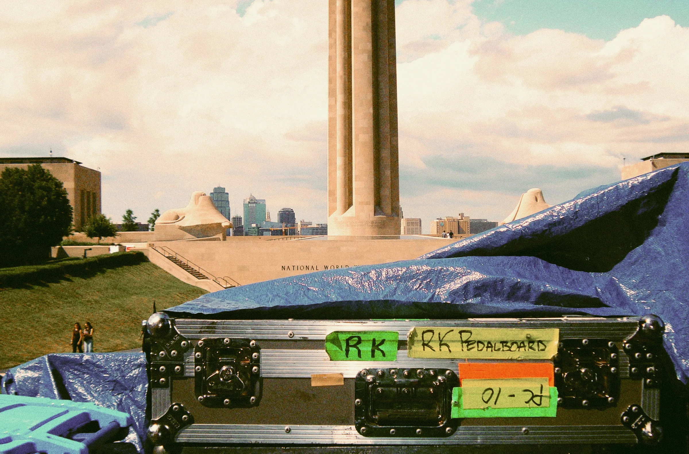 A road case covered in gaffer tape labels, parked on grass with the Liberty Memorial tower and the Kansas City skyline behind it