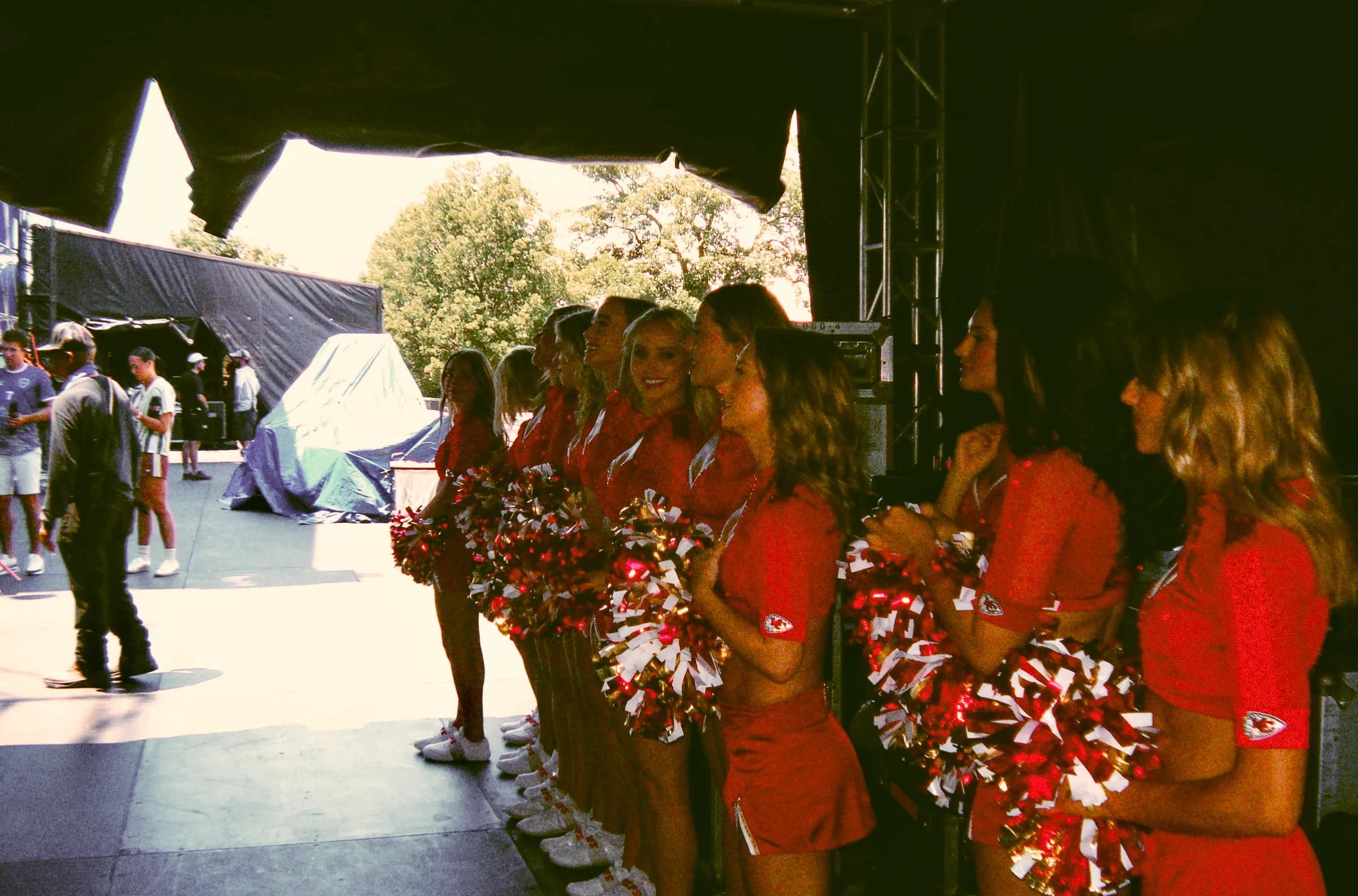 Cheerleaders in red waiting in the wings beside the stage, the festival ground stretching out behind them