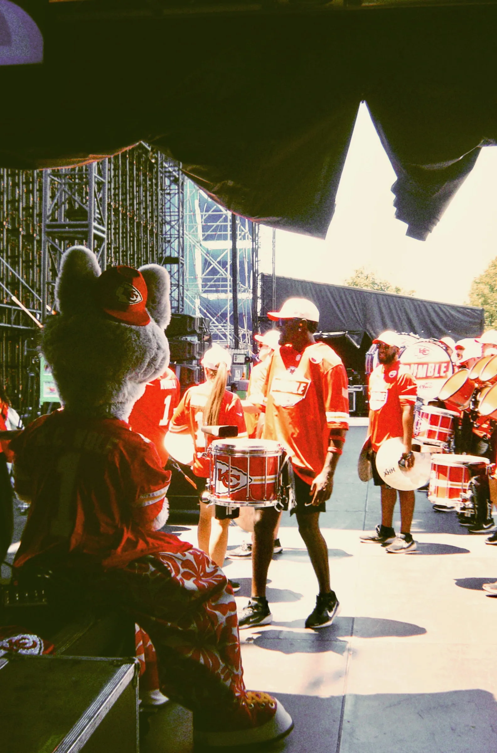 A costumed mascot watching a drumline perform, seen from behind the stage stacks