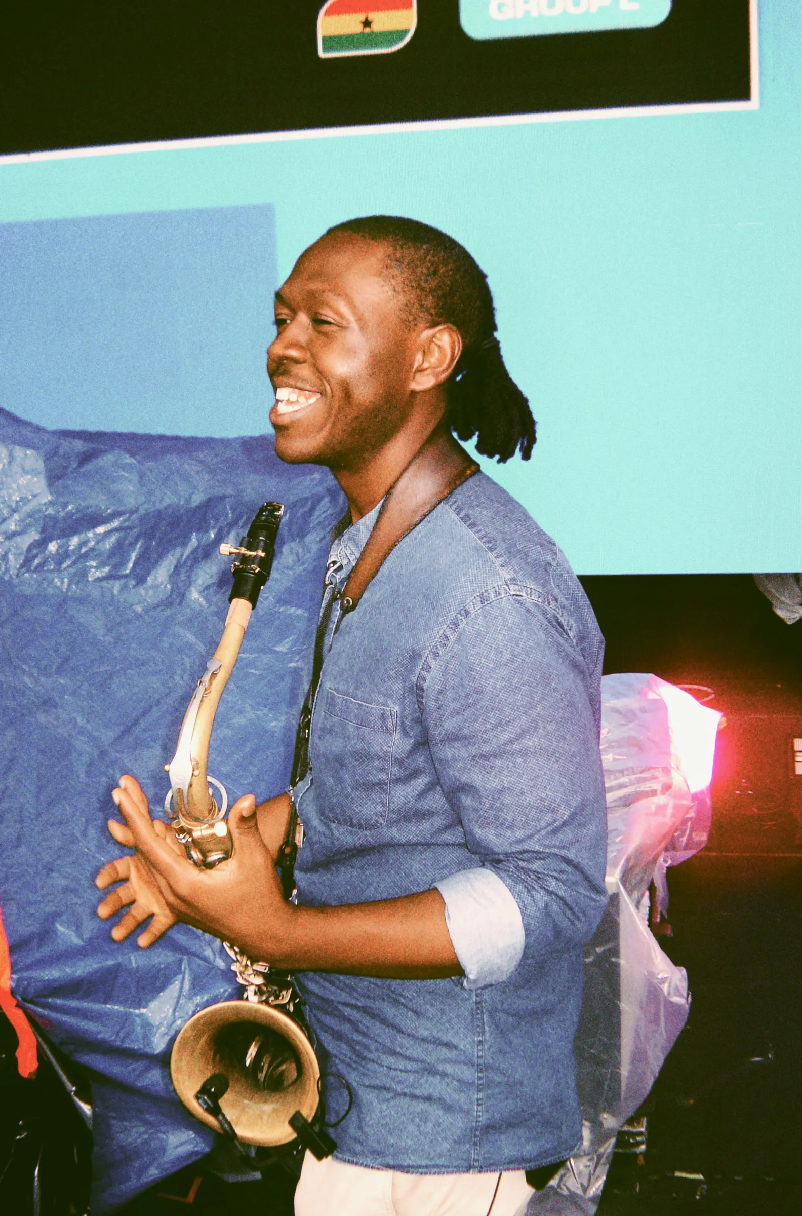 A saxophonist laughing mid-performance, lit against a bright teal screen