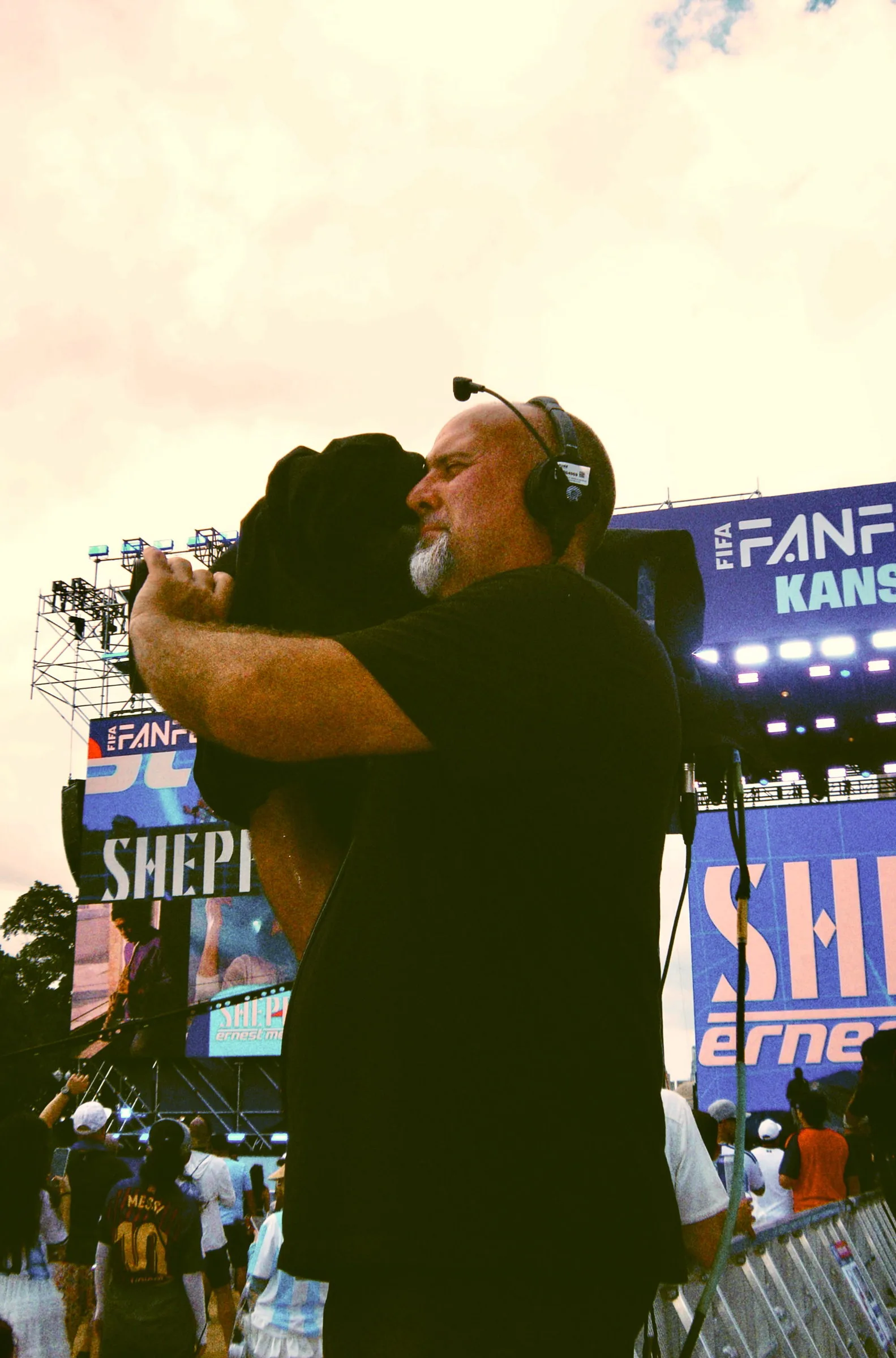 Two crew members in headsets holding each other in a long hug, festival screens glowing behind them