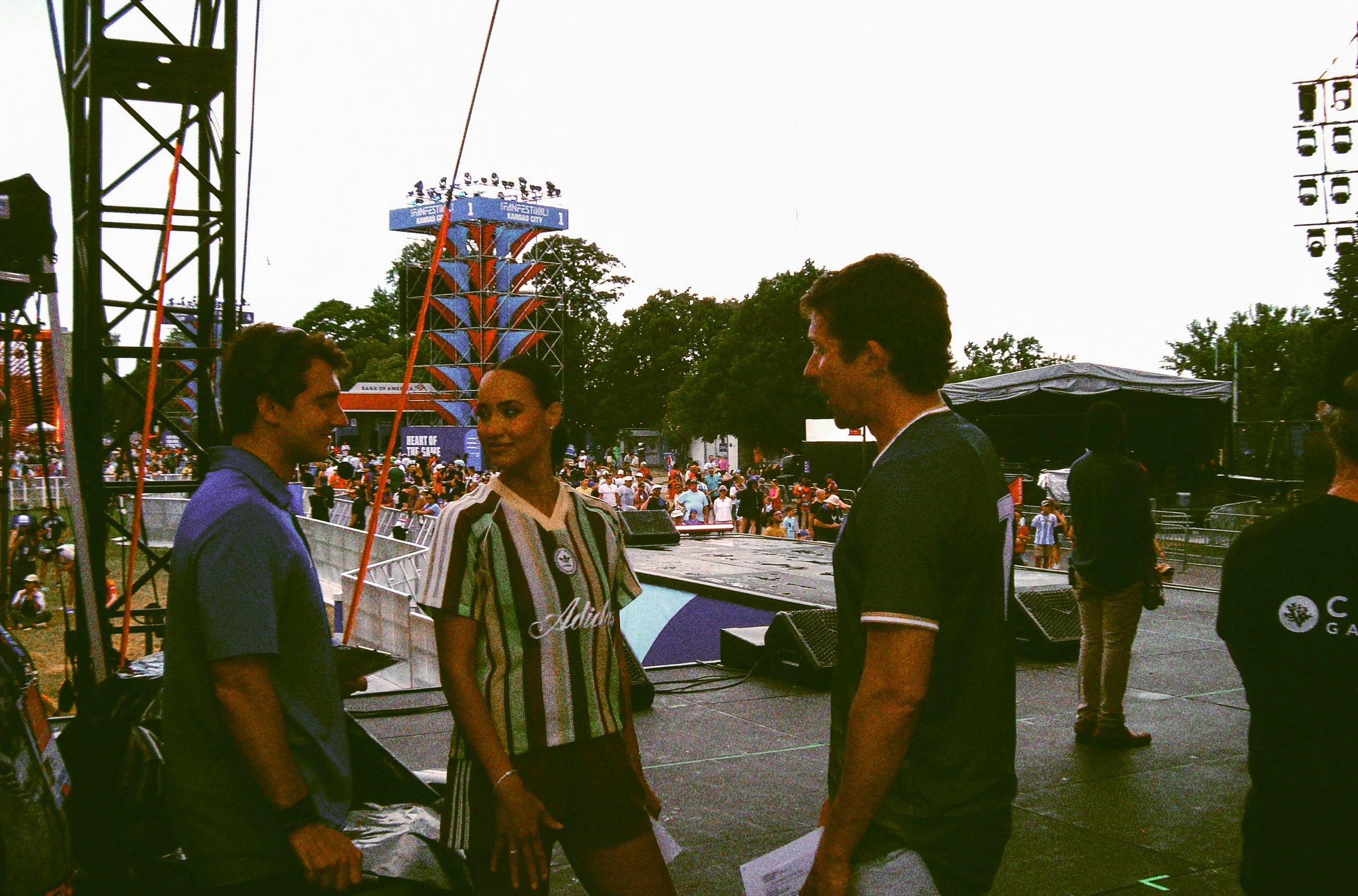 Three people talking on the ramp at the side of the stage as the crowd gathers beyond
